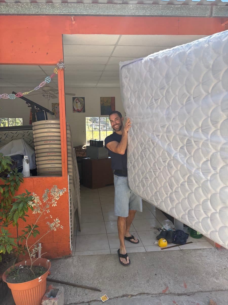 Smiling man holds a large white mattress wrapped in plastic outside an orange building.