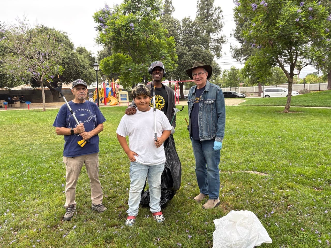 Four volunteers of different ages holding trash grabbers and bags in a grassy park.