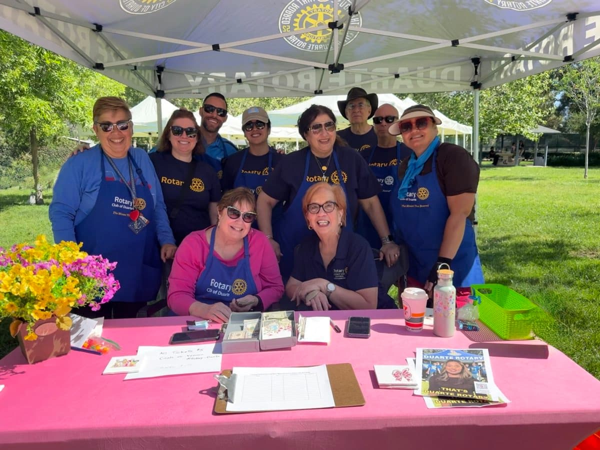 Smiling Rotary Club of Duarte members pose behind a pink table at an outdoor event.
