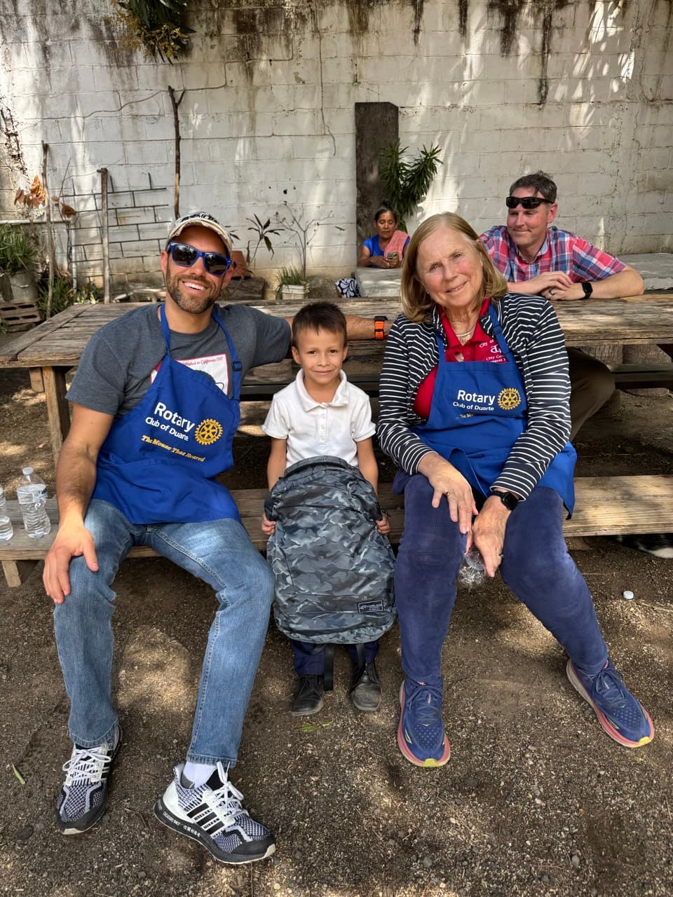 Two Rotary Club members in blue aprons pose with a boy holding a camouflage backpack.