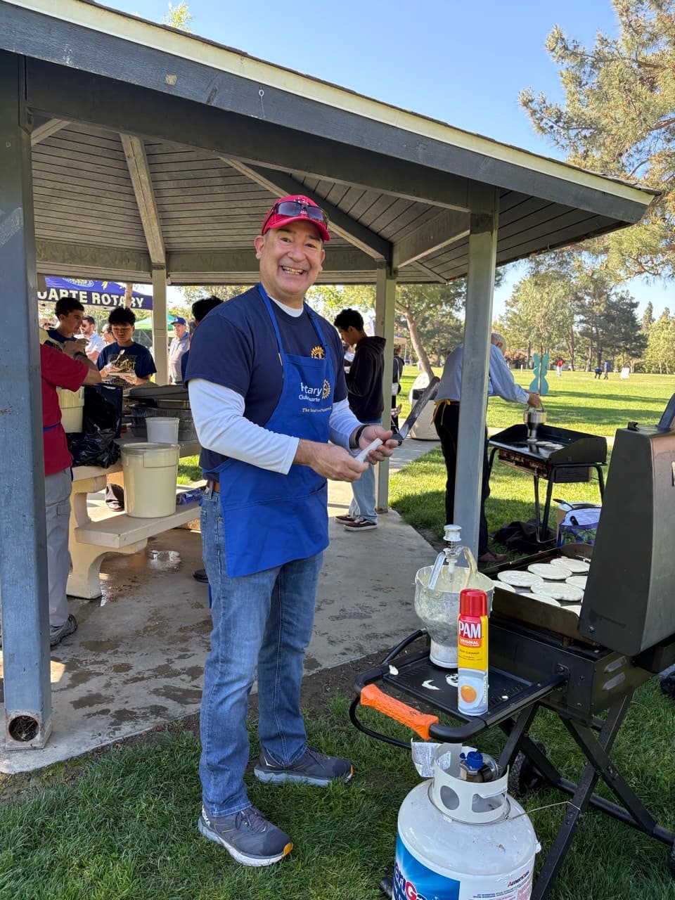 Smiling man in a blue apron cooks pancakes on a griddle under a park gazebo.